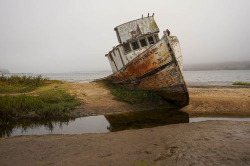 Free Weathered wooden boat beached on sand at Point Reyes with calm sea and cloudy sky. Stock Photo