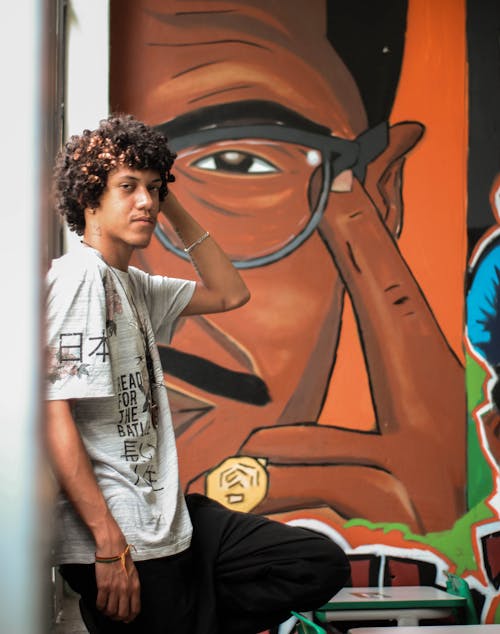 Side view of African American male with curly hair standing with hand on head near creative graffiti wall and looking at camera