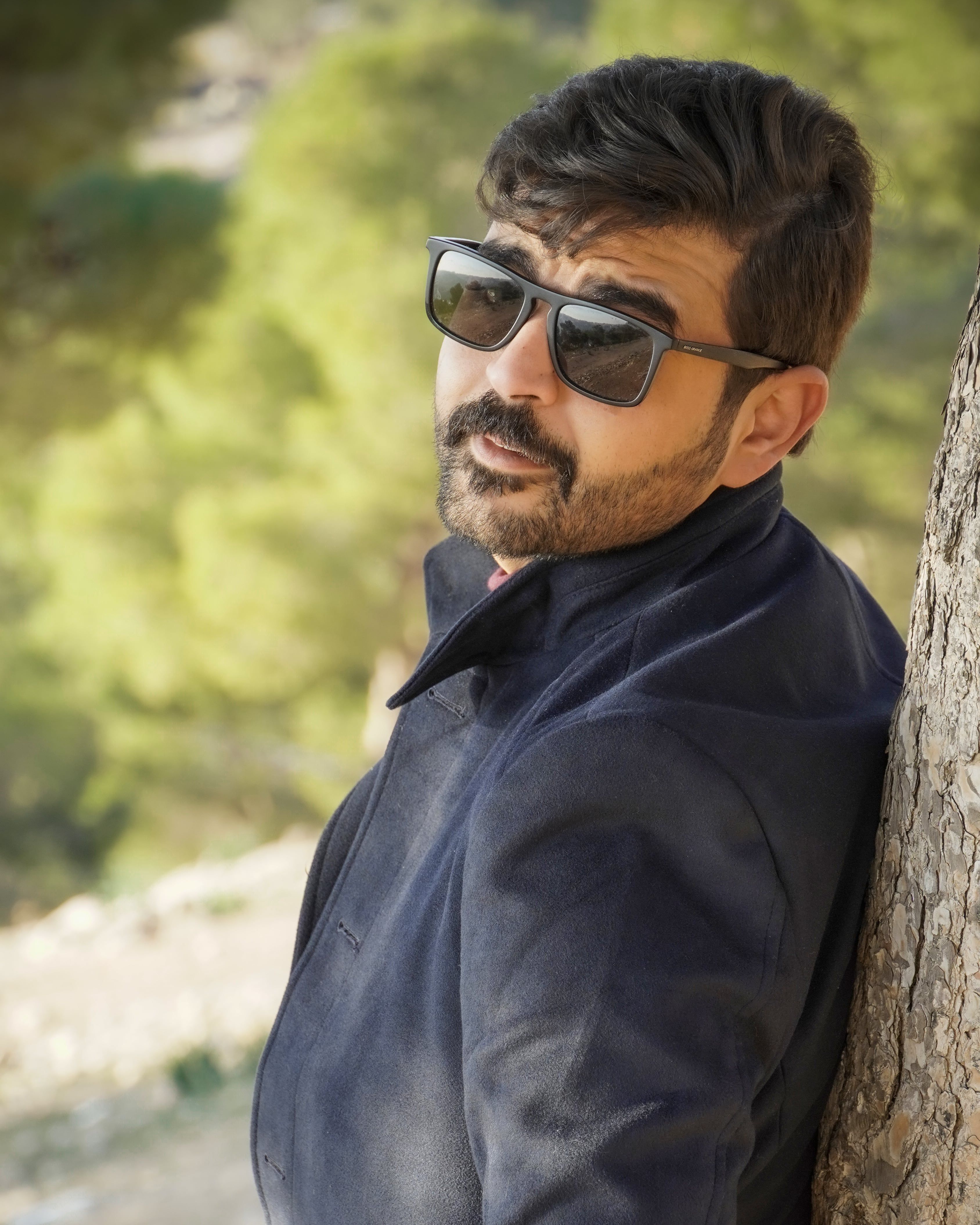 Stylish bearded man with sunglasses leaning on tree in sunny nature setting.