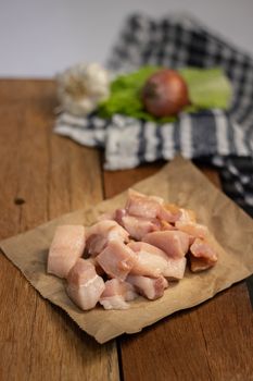 Chopped raw pork belly pieces on a brown paper over a wooden surface with vegetables.