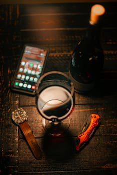 Aesthetic arrangement of wine glass, bottle, watch, phone, and knife on wooden table.