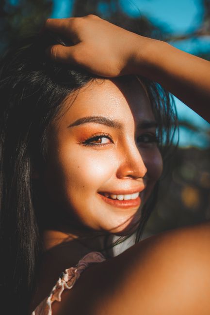 A close-up shot of a smiling woman outdoors, hand on head, warm lighting.
