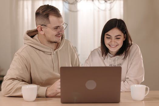A happy couple in hoodies working together on a laptop at home, enjoying hot drinks.