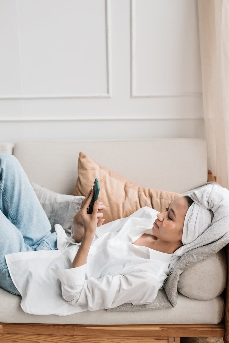 Woman With A Face Mask And Hair Wrapped In A Towel Lying On A Couch Scrolling Through Her Phone