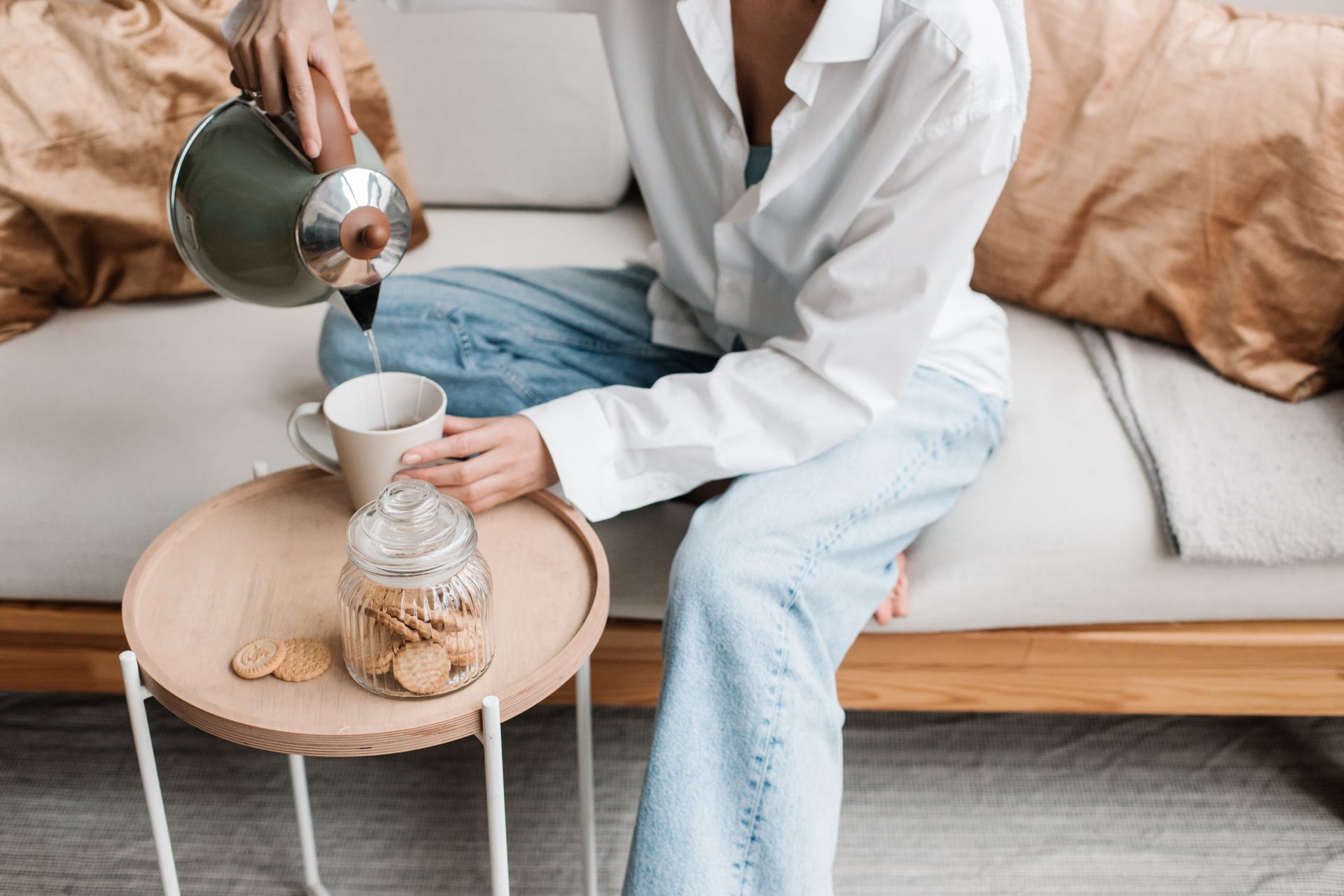 Person pouring hot drink into a cup next to cookies in a cozy living room setting.
