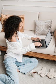 Woman typing on laptop while sitting on floor in cozy home interior with coffee.