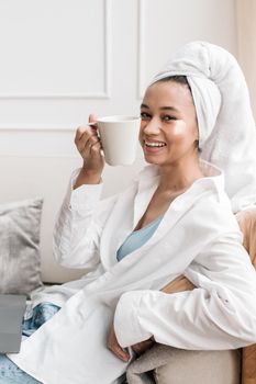 Happy African American woman enjoying a coffee at home, wrapped in a towel, exuding a relaxed and cozy vibe.
