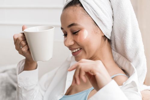 Free Woman with towel and eye patches enjoying coffee, embracing morning self-care routine. Stock Photo