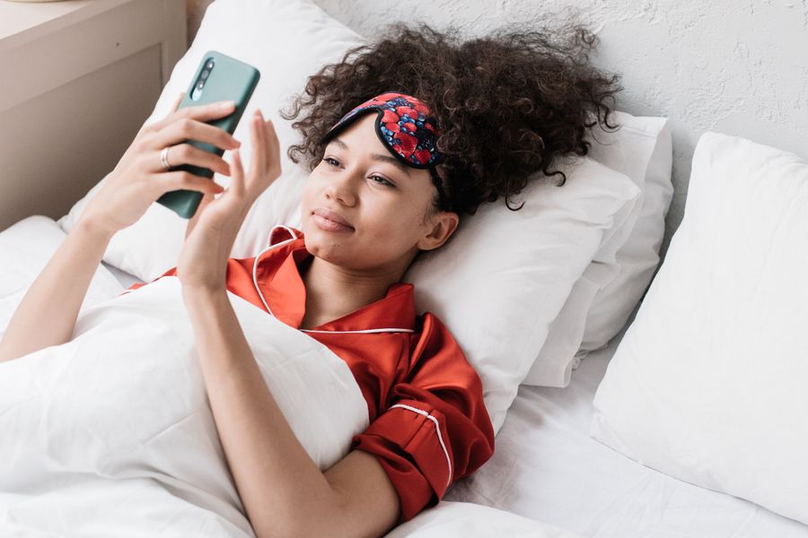A person in bed with a sleep mask on their forehead checking health data on a phone in the morning