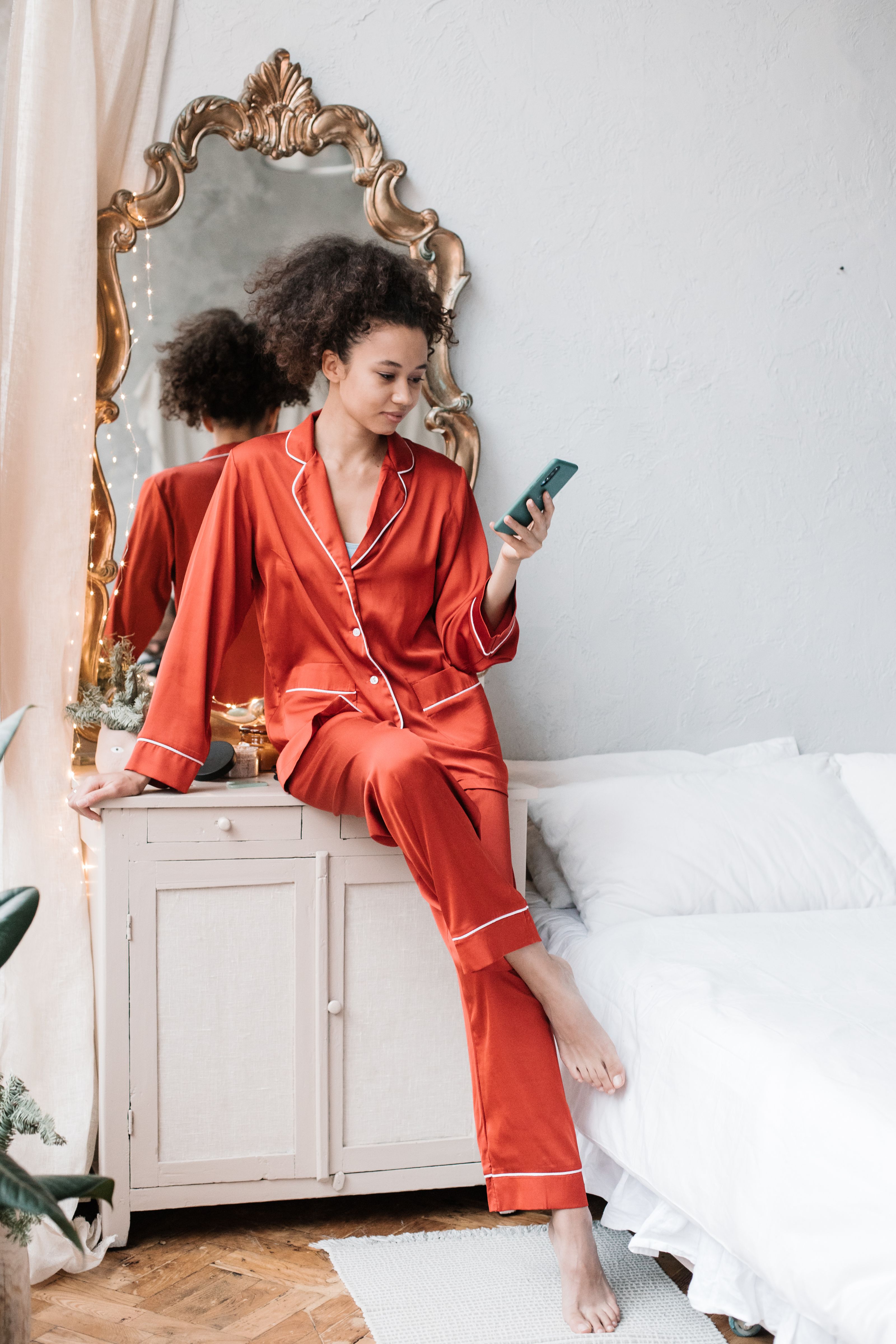 Mulher de pijama vermelho sentada na cômoda usando smartphone ao lado do espelho. Quarto brilhante e aconchegante.
