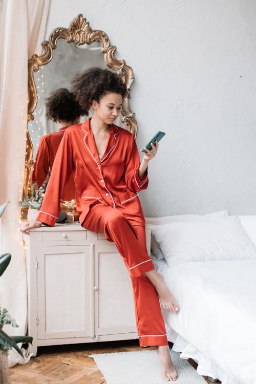 Free Woman in red pajamas sits on dresser using smartphone by mirror. Bright cozy bedroom. Stock Photo