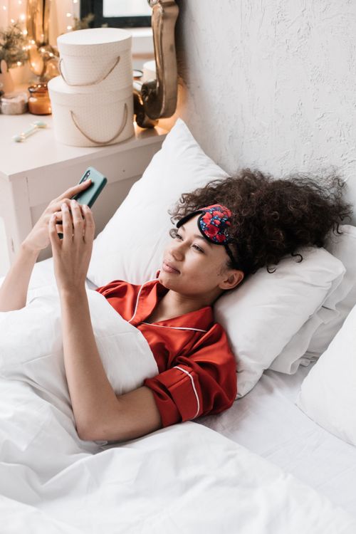 Free A young woman in red pajamas using a smartphone while lying in bed, wearing a colorful sleep mask. Stock Photo