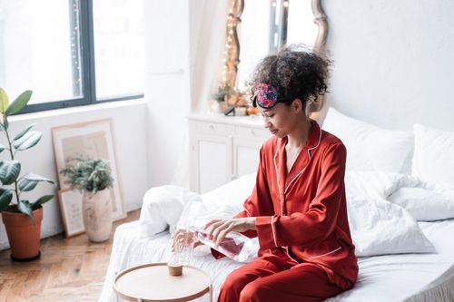 Free A woman in red pajamas pours water from a carafe into a glass, sitting on a bed in a cozy bedroom. Stock Photo