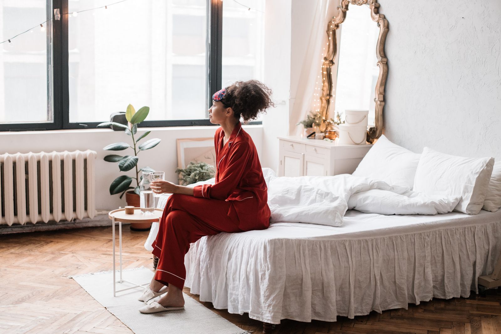 Woman in red pajamas sits on a white bed by tall windows, holding a glass of water