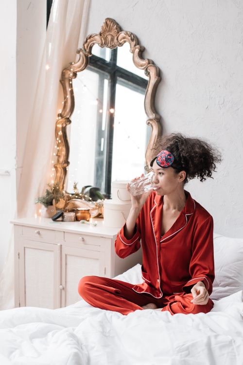 Free Woman in red pajamas sitting on bed, drinking water with decorative mirror backdrop. Stock Photo