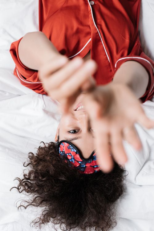 Free Smiling woman wearing a sleep mask reaches out playfully while lying on bed. Stock Photo