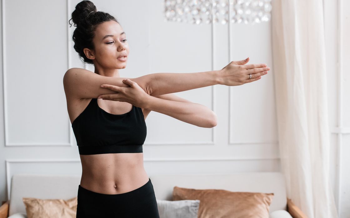Young woman in activewear doing an arm stretch indoors in a bright living room.