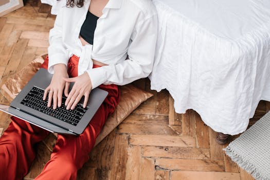 A woman in a white shirt and red pants typing on a laptop while seated on a pillow indoors.