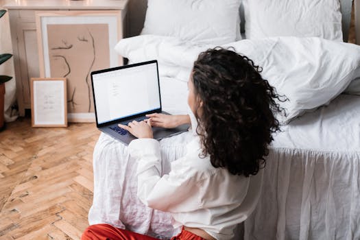 A woman works on her laptop while sitting on the bedroom floor, creating a comfortable home office environment.