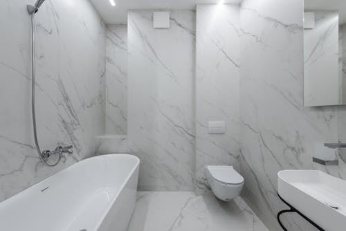 Interior of stylish bathroom with marble walls and bath placed near toilet and ceramic sink in light room with mirror