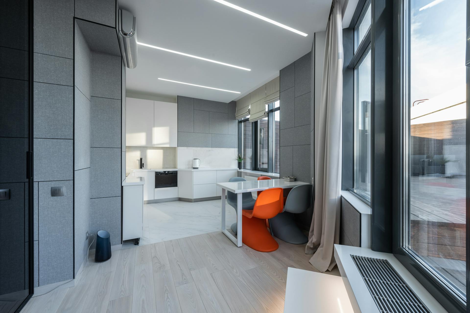 Interior of stylish light flat with table and colorful chairs placed near gray wall with windows against modern kitchen counter