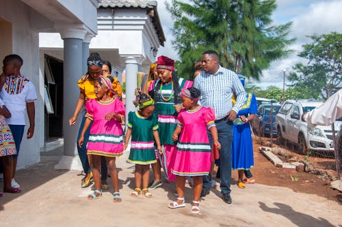 Cheerful African family in bright pink and green national dresses and elegant suit near house