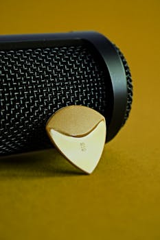 A close-up of a guitar pick resting against a microphone on a yellow background.