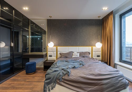 Modern light bedroom interior with soft bed with pillows and blanket near lamps and mirror with shelf near pouf and window with curtains