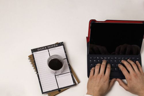 Free Top-down view of hands typing on a tablet, with coffee and a planner nearby. Stock Photo