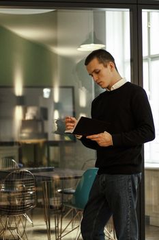 Caucasian man standing indoors, reading a notebook near a glass wall.