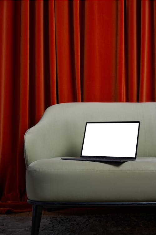 Laptop with white screen placed on soft couch on carpet near red curtains in bright room