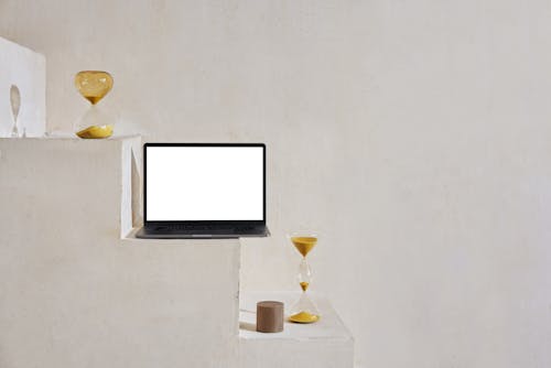 Netbook with white screen placed on white stone stairs near sand glasses and small round figure near wall in bright room