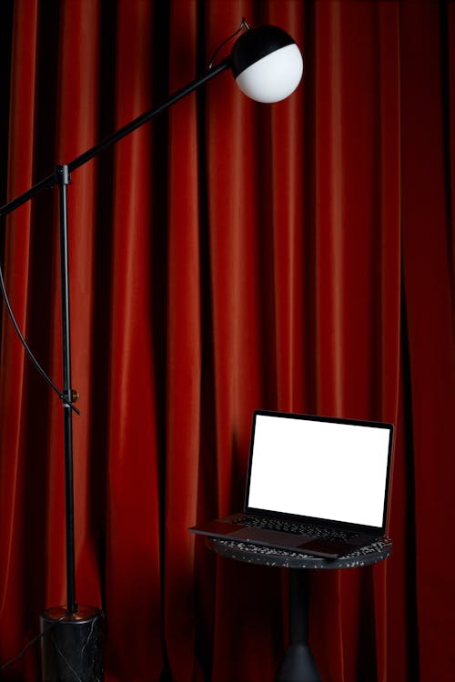 Modern netbook with white screen placed on round table near tall lamp and red curtains in light room