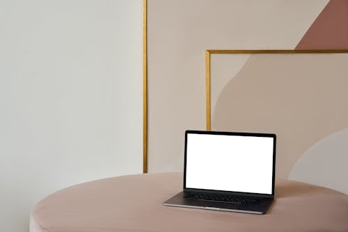 Free Laptop with white screen placed on soft beige pouf near framed paintings and white wall in light room Stock Photo
