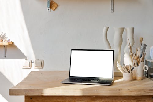 Netbook with white screen placed on wooden table near uneven curved ceramic vase near holders with brushes and tools near shelf in light workshop with white wall