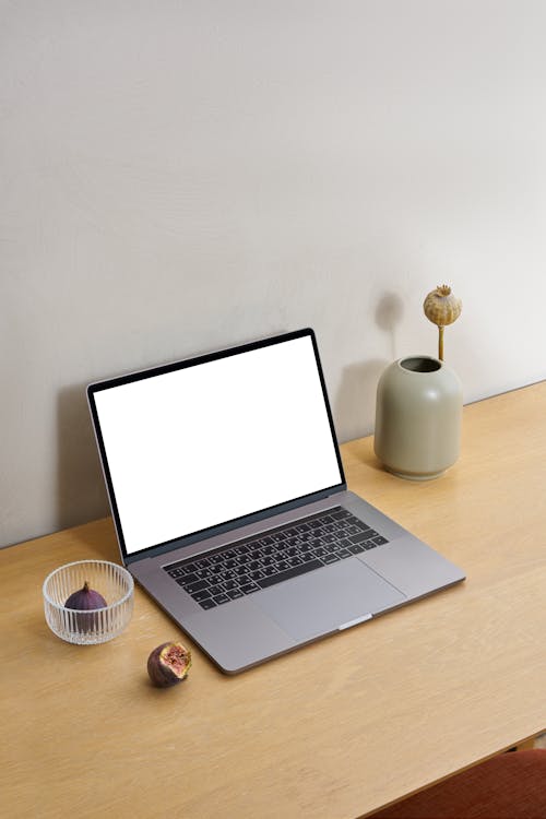 Laptop with white screen placed on wooden table near glass bowl with figs and ceramic vase near wall in bright room