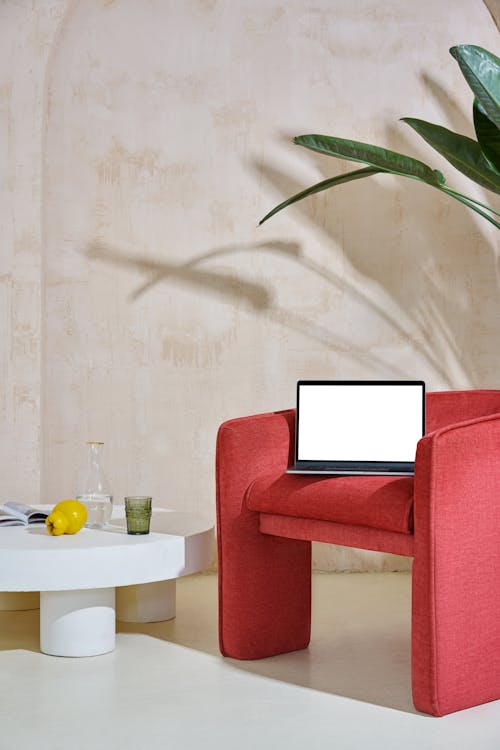 Laptop with white screen on red armchair near small white table with glass near jug with water and apple with magazine and plant with green leaves near wall in room