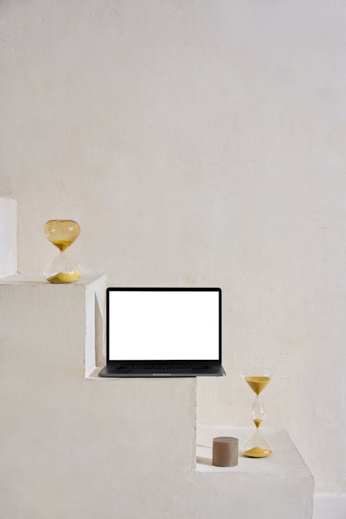 Modern netbook with white screen on stone stairs near sand glasses and small round figure near white wall in bright place