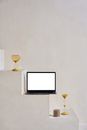 Modern netbook with white screen on stone stairs near sand glasses and small round figure near white wall in bright place