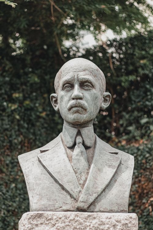 Free Stone bust of a mustached man in a suit and tie, surrounded by greenery. Stock Photo