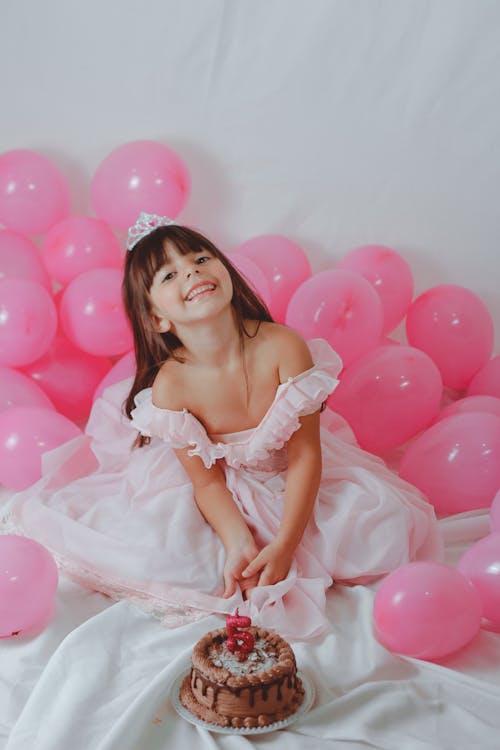 Free From above of positive little girl in costume of princess near sweet delicious birthday cake among pink balloons Stock Photo