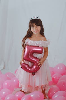 Full body of happy adorable little girl in diadem smiling among shiny pink balloons