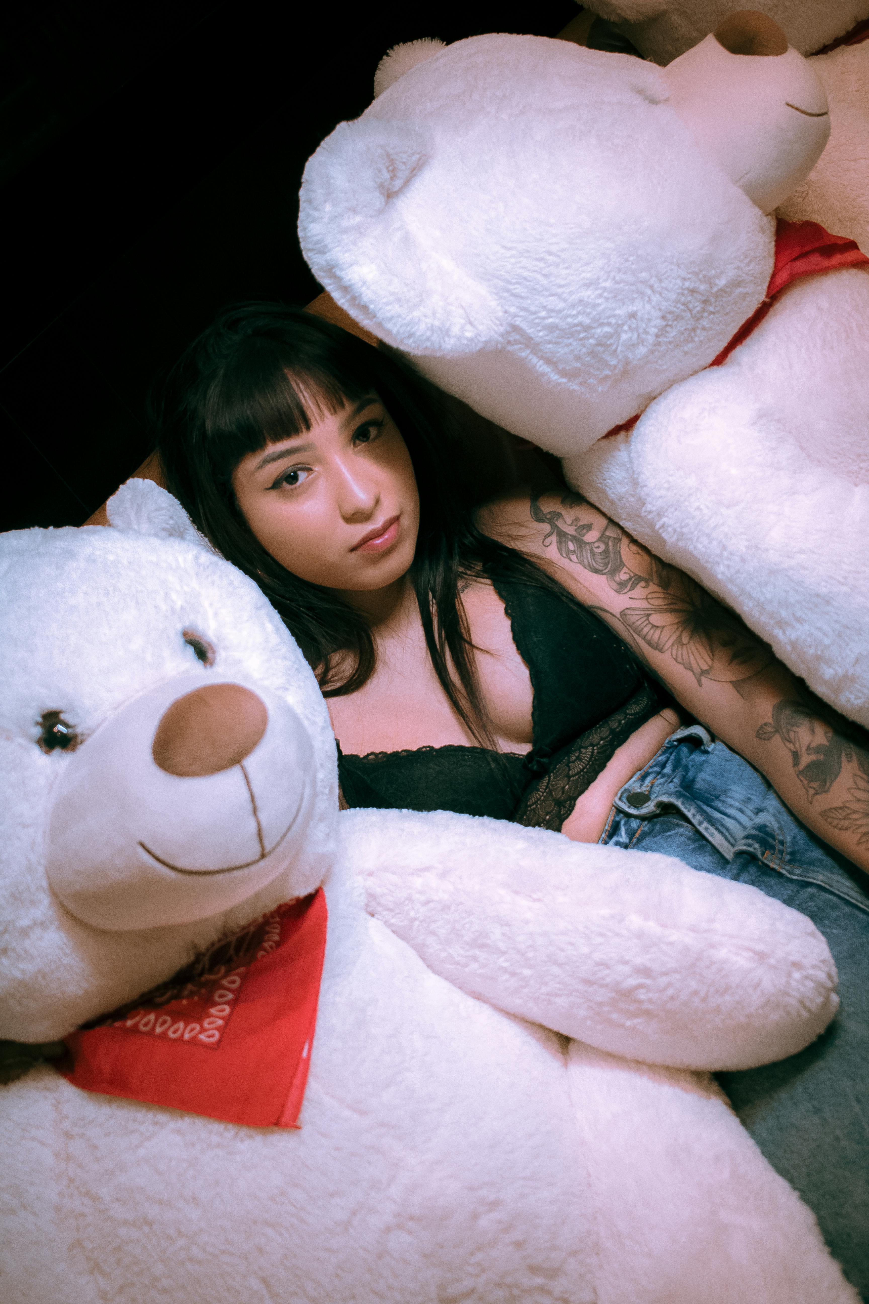 A tattooed woman in casual attire reclines among large white teddy bears in a cozy, indoor setting.