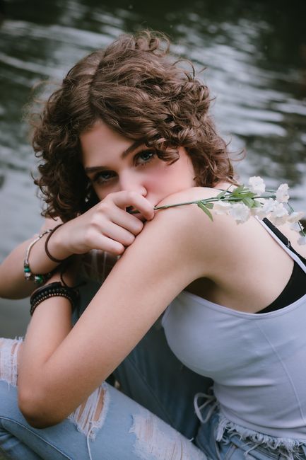 Side view of romantic young lady with curly hair in casual outfit resting on shore of lake and looking at camera with stem of delicate flowers in hand