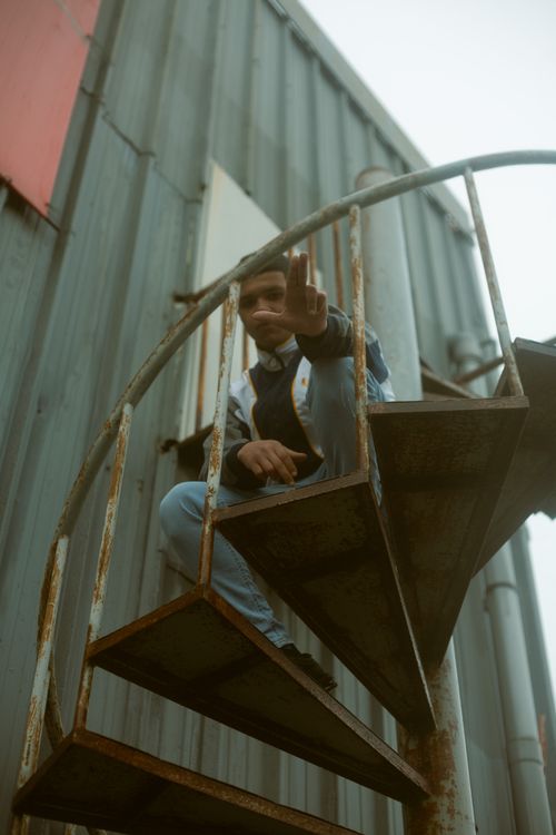 Free A young man sits casually on a rusty spiral staircase outdoors, giving a relaxed atmosphere. Stock Photo