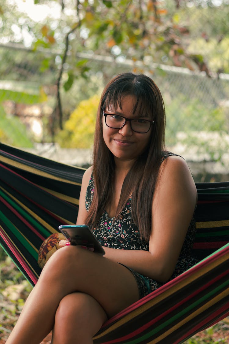 A Woman With Eyeglasses Sitting On A Hammock While Holding A Mobile Phone