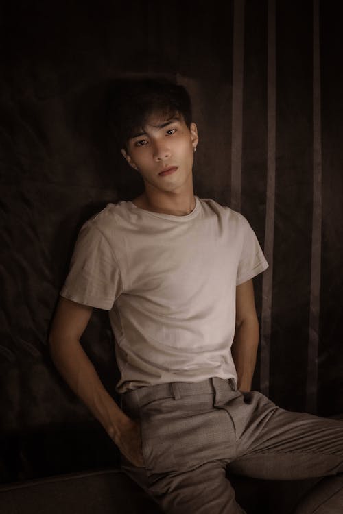 Free Confident Asian male in casual clothes sitting with hands in pockets and looking at camera in dark room Stock Photo
