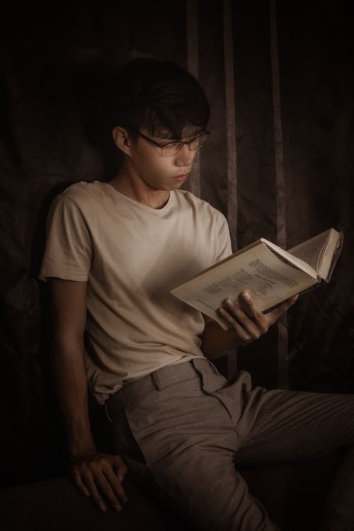 Free Thoughtful Asian male in eyeglasses and casual clothes sitting and reading book in dark room Stock Photo
