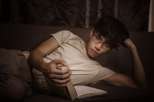 Free Young serious Asian guy in casual outfit and eyeglasses lying on sofa while leaning on hand while reading interesting novel in light room Stock Photo