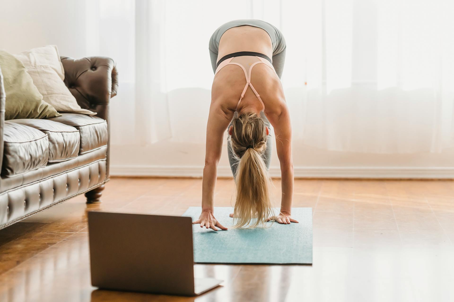 Full body of unrecognizable female in activewear performing standing forward bend asana while standing on mat near laptop during online yoga lesson at home